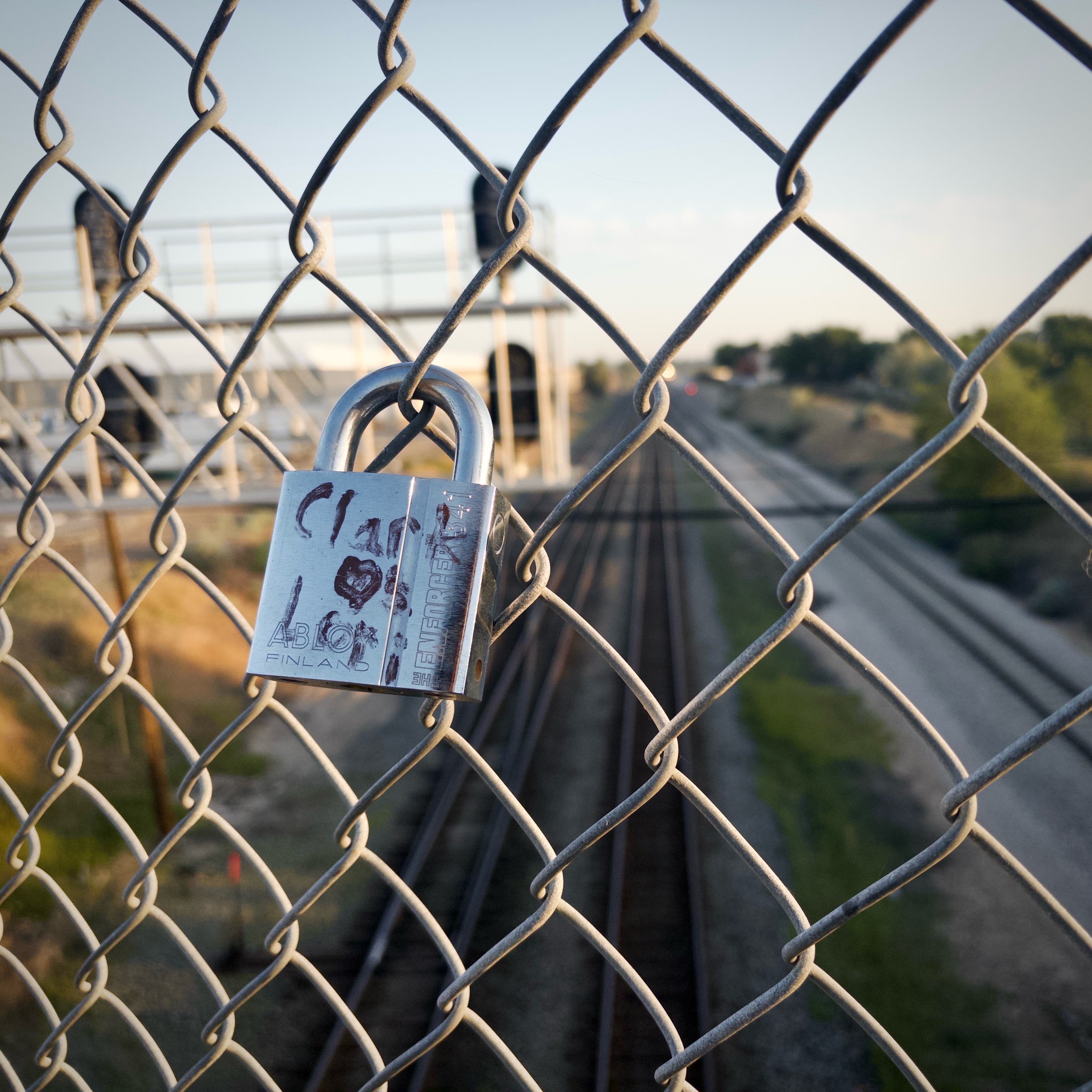 A lock on a chain link fence overlooking some train tracks. Written on the lock in paint marker is “Clark hearts Lori.”